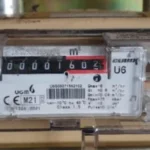 a gas meter that is housed in a brown casing, it is attached to some pipes at the top.