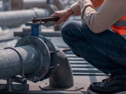 a man who's wearing a orange high vis jacked and is crouched down in order to fix a heat pipe with a spanner.