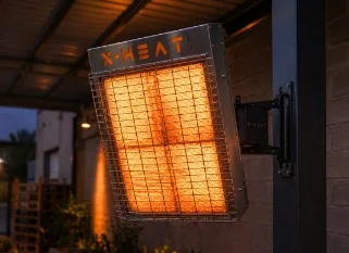 a wall mounted heater that is attached to a cuboid shaped metal pillar via a bracket that then attaches to the heater itself. The heater has 4 big vents on the front of it, each of them is glowing orange because it's turned on, the heater and pillar are black. Above the ventilation there is the orange all caps X-HEAT logo.. The heater is in a warehouse.