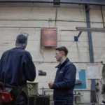 two workers standing underneath a x-heat heater that is turned on.